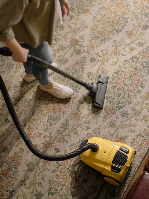A child wearing beige shoes, blue jeans, and a beige jacket operates a yellow and black vacuum cleaner with a hose attachment on a patterned area rug in a room with hardwood flooring, during a surface cleaning session. The rug features intricate floral designs in shades of cream, green, and orange. The lighting is natural and soft, highlighting the clean and maintained condition of the rug. This scene exemplifies domestic cleaning practices, with Hackney Carpet Cleaning providing deep cleaning and sanitisation services to maintain hygiene in home interiors.