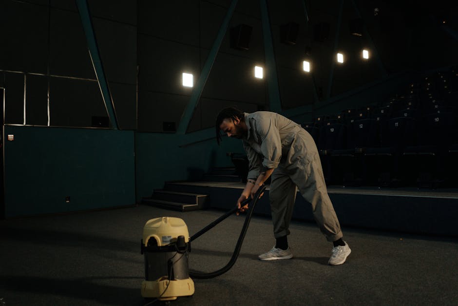 A professional in full protective gear, including a white hazmat suit, face mask, and blue gloves, is performing deep cleaning or sanitisation on a beige carpet in a residential living room. The technician is using a carpet cleaning machine that releases steam or cleaning solution, creating visible white vapor on the carpet surface. The room is well-lit with natural light, featuring light-colored walls, a wooden bookshelf with decorative items, and several potted plants. This image illustrates effective surface cleaning and sanitisation processes performed by Hackney Carpet Cleaning to maintain hygiene and cleanliness in domestic spaces, aligning with their service of deep cleaning and carpet sanitation showcased in the Dalston guide to emergency carpet cleaning E8, Hackney.
