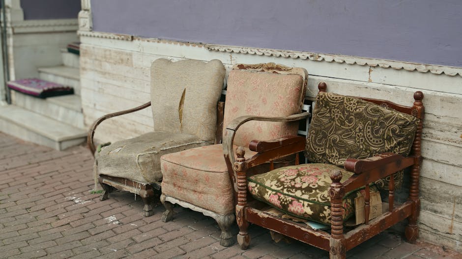 Three old and worn-out upholstered armchairs with peeling fabric and broken frames are placed on a brick sidewalk beside a weathered white wooden wall with peeling paint. The first armchair on the left has a faded beige fabric with torn upholstery and missing cushioning. The middle chair features pinkish, patterned fabric that is heavily frayed and stained, with parts of the fabric detached from the frame. The rightmost chair has a dark wood frame and is upholstered with a green and pink floral patterned fabric, which shows signs of age, wear, and some exposed padding. Behind the chairs, a set of concrete steps with a doormat at the base is visible, indicating an entrance to a building. The scene suggests outdoor furniture in need of cleaning, restoration, or proper disposal, consistent with waste management policies like those outlined by Hackney Council for upholstery cleaning waste. Hackney Carpet Cleaning specializes in surface and deep cleaning services to maintain hygiene and restore furniture cleanliness.
