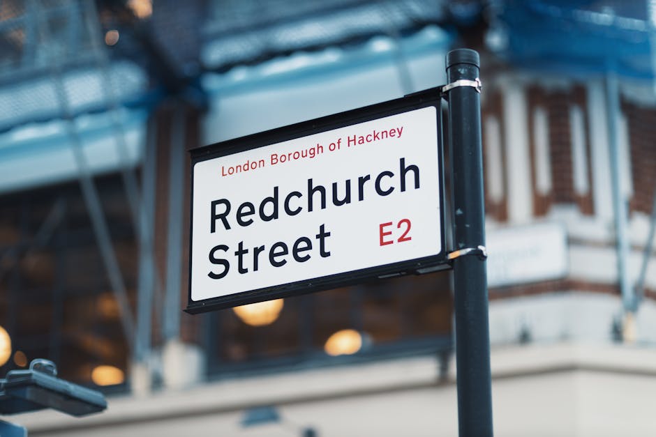 Close-up view of a street sign mounted on a black metal pole indicating 'Redchurch Street' in the London Borough of Hackney, with the postal code E2 in red. The sign is on a busy urban street with blurred commercial buildings in the background, which feature brick facades, large windows, and some scaffolding or construction elements. The lighting suggests daytime, with natural light illuminating the sign and surroundings. This street signage is part of an area that may require professional surface cleaning and maintenance, as offered by Hackney Carpet Cleaning, to ensure cleanliness and hygiene in both residential and commercial properties.