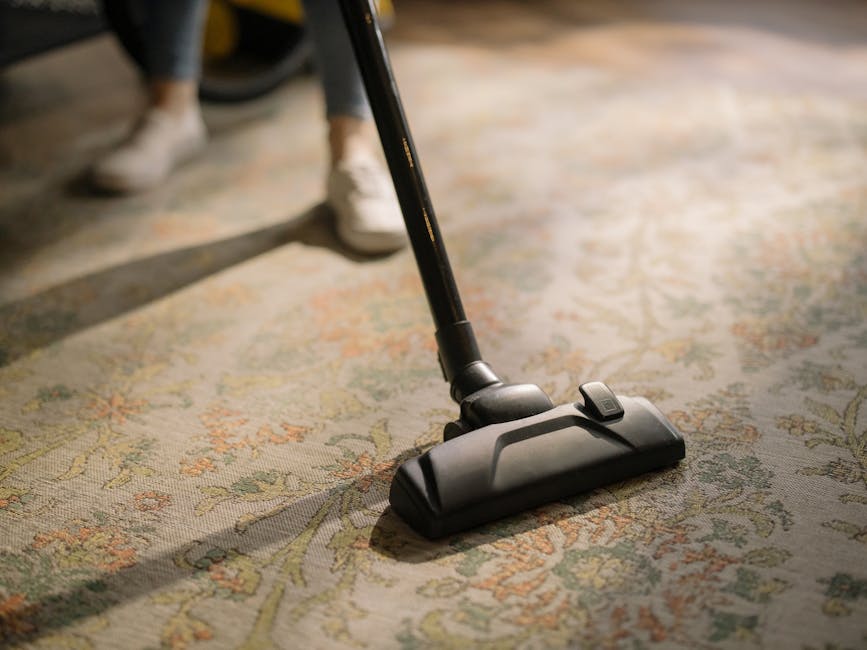 Close-up of a person vacuuming a patterned oriental rug in a residential living room, with visible feet in white sneakers and part of a body in casual clothing, demonstrating surface cleaning and deep cleaning for hygiene. The vacuum cleaner's head is black and sleek, positioned on the richly colored rug featuring floral motifs in shades of beige, green, and orange, while the background shows a wooden floor and some furniture, illuminated by natural light. This image illustrates professional carpet and floor cleaning techniques, emphasizing cleanliness and maintenance as part of Hackney Carpet Cleaning's domestic cleaning services in London Fields.