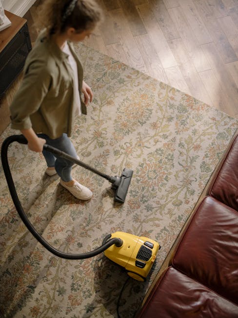 A woman with curly hair, wearing a beige jacket, blue jeans, and white sneakers, engaged in deep cleaning a patterned carpet with a yellow vacuum cleaner in a living room. The room features wooden flooring partially covered by the carpet, a brown leather sofa, and a wooden entertainment unit on a wall painted in a light neutral shade. Natural light illuminates the space, highlighting the cleanliness and detailed surface cleaning process. Hackney Carpet Cleaning's service is focused on comprehensive carpet hygiene and surface sanitisation, demonstrating professional domestic cleaning methods. The scene emphasizes precise surface cleaning and maintenance in a residential setting.