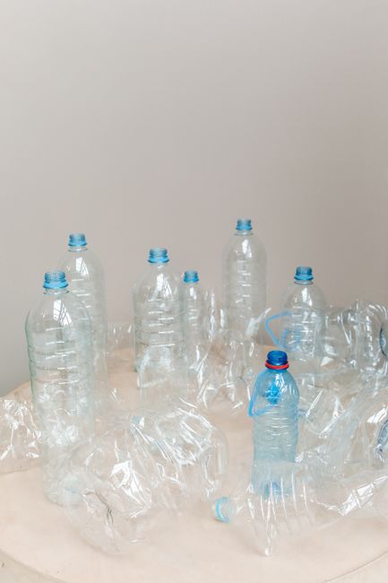 A collection of empty, crushed plastic water bottles with blue caps scattered on a light-colored surface against a plain light gray wall. Some bottles are upright, while others are flattened or leaning. The scene demonstrates plastic waste awaiting responsible recycling or disposal. The lighting is even and natural, emphasizing the transparency and crumpled textures of the bottles. This image reflects proper waste management practices associated with professional cleaning services like Hackney Carpet Cleaning, emphasizing cleanliness and environmental responsibility in the context of recycling after cleaning.