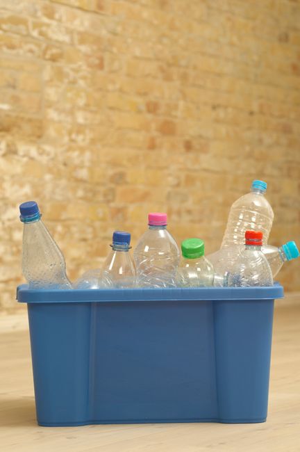 A blue plastic recycling bin filled with several empty plastic bottles of various sizes, some with colorful caps such as blue, green, pink, and red. The bin is placed on a light wooden floor in front of a textured, yellowish brick wall. The bottles are clear, and some are slightly crumpled, indicating they have been used. The scene suggests responsible disposal of plastic waste after cleaning or recycling, aligning with the theme of surface cleaning and maintenance at Hackney Carpet Cleaning, with a focus on eco-friendly practices in domestic or commercial settings.