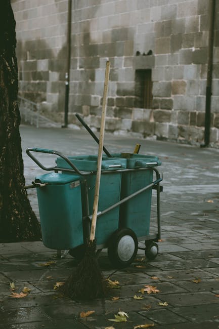 A cleaning cart with a large teal plastic bin and attached smaller containers is situated on a paved sidewalk next to a tree, with a broom leaning against it. The sidewalk appears wet, possibly from recent cleaning, with scattered fallen leaves around. The background features a stone wall with a small, dark opening and a black pole nearby. The scene is outdoors in natural lighting, emphasizing the maintenance tools used for surface cleaning in a city environment. The presence of the cart and broom indicates active cleaning efforts, consistent with professional cleaning services like Hackney Carpet Cleaning, addressing hygiene and surface maintenance in urban areas.