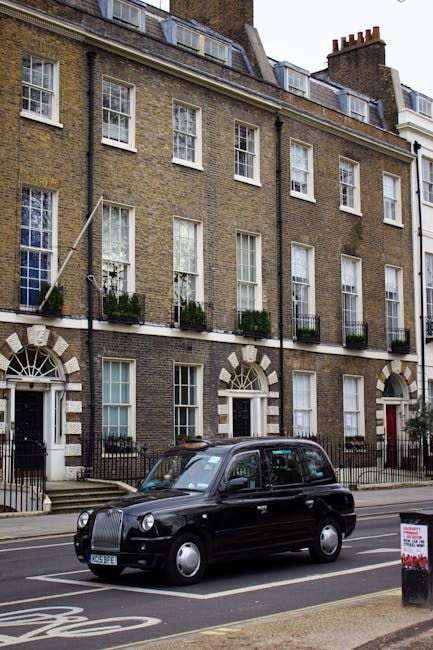 Image of a dark green London taxi parked on a street in front of a multi-story brick residential building with white-framed sash windows and small balconies adorned with potted plants and flowers. The building's facade features decorative stone archways above the ground-floor entrance. The street is paved, with a sidewalk and a black and white signpost visible on the right side of the image. The scene is illuminated by natural daylight, highlighting the cleanliness of the surroundings. Hackney Carpet Cleaning emphasizes the importance of surface cleaning and maintenance in its services, ensuring domestic spaces remain hygienic and well-maintained, aligning with the insights in 'What to know about hidden cleaning charges in Hackney, HACKNEY.'